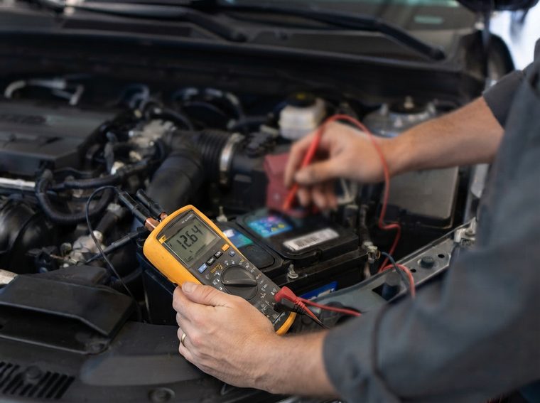 Mains d’un mécanicien mesurant la tension d’une batterie de voiture au multimètre, pointe rouge sur la borne positive, afficheur à 12,64 V
